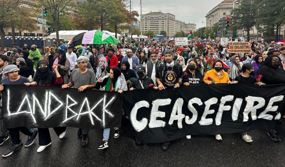 Thousands of people march in the streets of Washington, D.C. holding signs. Two banners in the foreground read LANDBACK and CEASEFIRE.