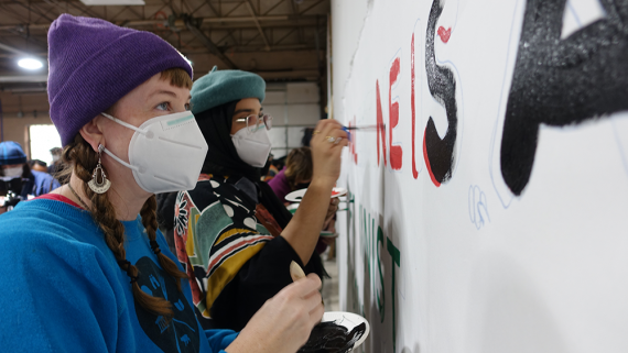 Two people wearing face masks paint a banner that says PALESTINE IS A FEMINIST ISSUE