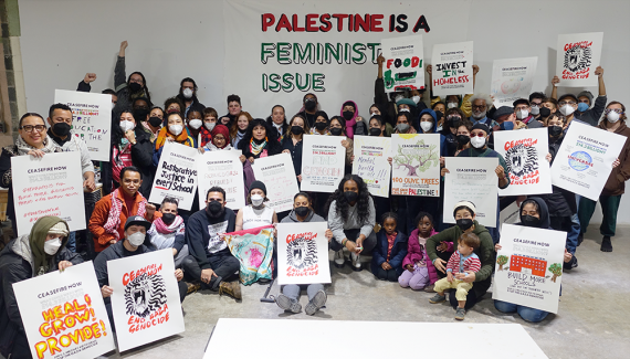 A large group of diverse people gather in front of a banner and hold signs supporting a ceasefire. Banner text: PALESTINE IS A FEMINIST ISSUE