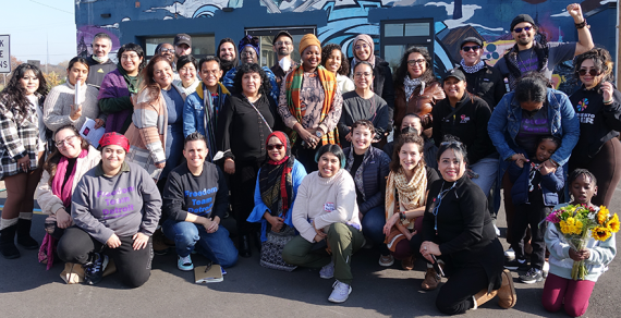 About 30 members group together outside a mural-covered building on a sunny day.