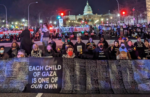 Hundreds of protesters holding many banners block a street in Washington, DC. at night, with the Capitol building in the background