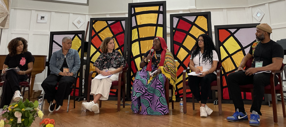 Photograph of six people sitting on a stage in front of decorative panels. The person in the center speaks into a microphone