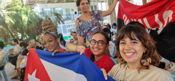 Photograph shows four people in the foreground holding banners and flags, seated outdoors along with many other people in the background