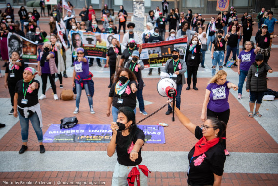 Photograph shows multiple people in formation with banners and pointing forward as part of a chant. The two people in front also have a bullhorn