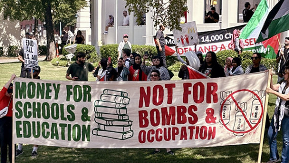 Dozens of people begin gathering with banners and flags in a grassy area. Front banner text: Money for Schools & Education / Not for Bombs & Genocide