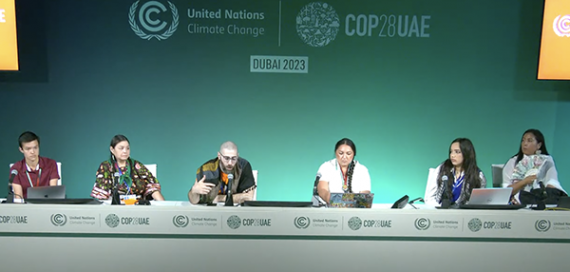 Six panelists sit at a long table in front of a green wall with COP28 graphics, speaking to audience members who are out of frame.
