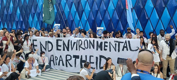 Photo of about 50 protesters, with others outside the photo frame, gathered around a large banner with the words “END ENVIRONMENTAL APARTHEID” in front of a blue rhombus-patterned wall.