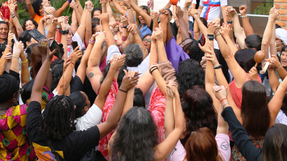 Dozens of people group together tightly outdoors, clasping each others’ raised hands.