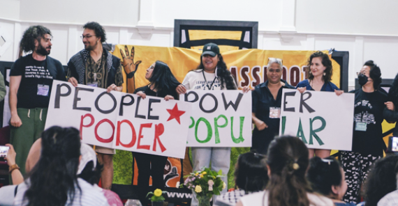 Seven people on stage hold signs while audience members observe. Sign text: PEOPLE POWER / PODER POPULAR