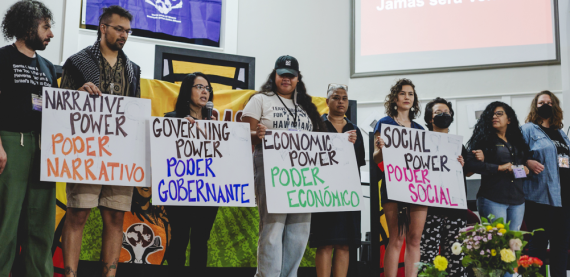 Nine people standing in a row holding four signs that read: NARRATIVE POWER/PODER NARRATIVO, GOVERNING POWER/PODER GOBERNANTE, ECONOMIC POWER/PODER ECONÓMICO, SOCIAL POWER/PODER SOCIAL.