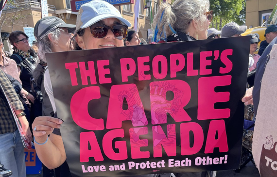 A GGJ member in sunglasses and a hat holds a sign at a march. Sign text: The People’s Care Agenda / Love and Protect Each Other!
