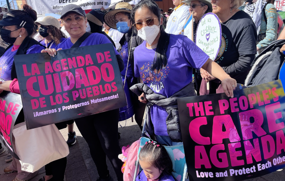 Photo of people in a march holding black and magenta signs in both English and Spanish. Sign text: THE PEOPLE’S CARE AGENDA / Love and Protect Each Other!