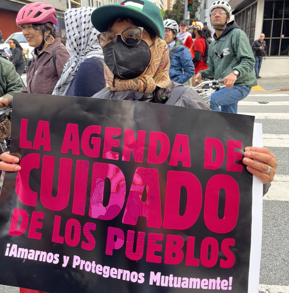 Un miembro de GGJ con bufanda y sombrero sostiene un cartel en una marcha. Texto del cartel: “La Agenda de Cuidado de los Pueblos / ¡Amarnos y Protegernos Mutuamente!