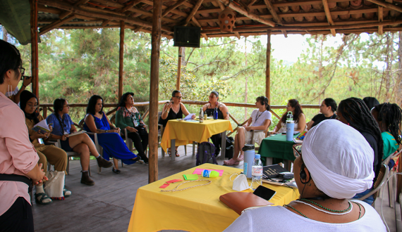 More than a dozen people sit in a large circle with some small tables, under a wooden pavilion with many trees in the background.