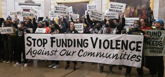 Dozens of people protest in the Capitol Building Rotunda, holding signs saying “DIVEST FROM MILITARISM, INVEST IN LIFE” and “CEASEFIRE,” along with a large banner with the words “STOP FUNDING VIOLENCE / Against Our Communities” 