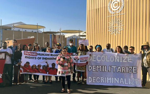 Delegation members holding banners with text: DEFEND HUMAN RIGHTS AND THE RIGHTS OF INDIGENOUS PEOPLES, DECOLONIZE, DEMILITARIZE, DECRIMINALIZE