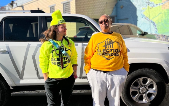 Two people stand outside a building on a sunny day, wearing Election Defender sweatshirts. A vehicle in the background has the word VOTE written on the doors in gray tape.