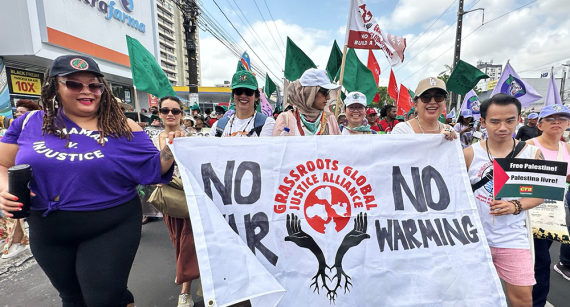 Photo of GGJ delegation members marching in Brazil holding a large white banner. The banner includes the GGJ logo with text: No War, No Warming. A small hand-held sign features the Palestinian flag with this text: Free Palestine! Palestina livre!