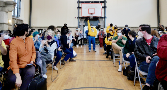 People sitting in rows of a packed auditorium look down the middle aisle to the back of the room, where other folks are standing, including one person with their fist in the air.