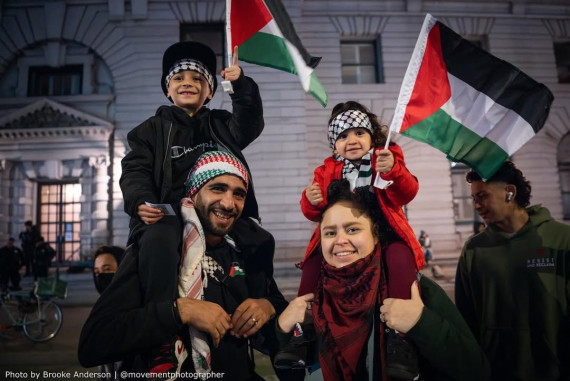 Two adults and two children smiling, with the children waving Palestinian flags and sitting on the adults’ shoulders